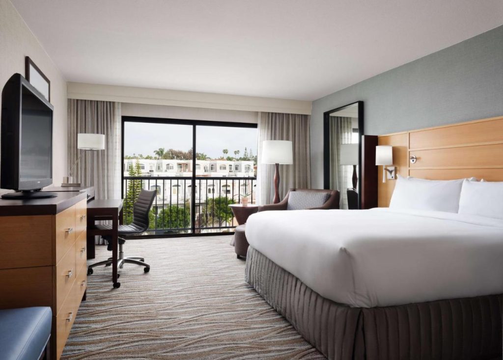 Nette hotelkamer met een groot bed, een bureau en een balkon met openslaande deuren en uitzicht op palmbomen en gebouwen