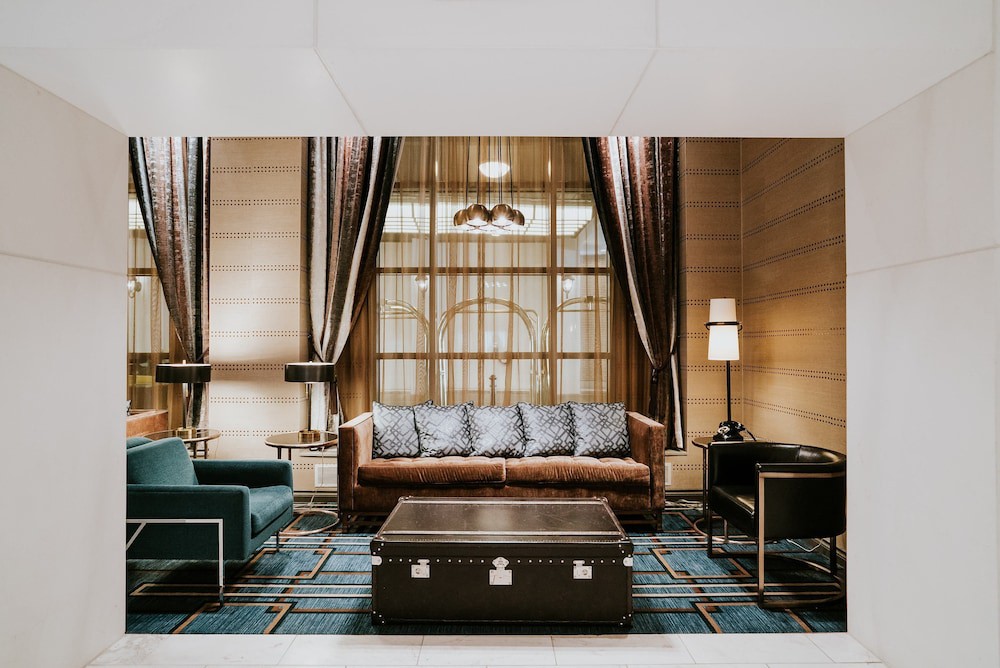 Een sfeervolle hotellounge met een bruine leren bank, een groene fauteuil en lange gestreepte gordijnen bij hoge ramen