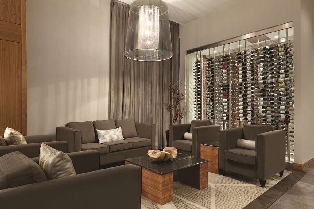 Lounge met grijze bankstellen rond een houten salontafel, een grote hanglamp en een glazen wand gevuld met wijnflessen op de achtergrond