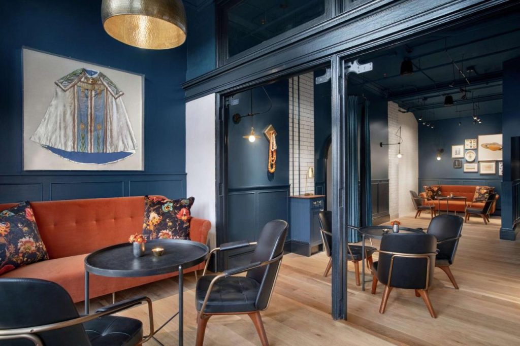 Loungeruimte met donkerblauwe wanden, een oranje bank met bloemenkussens en zwarte stoelen rond ronde tafels