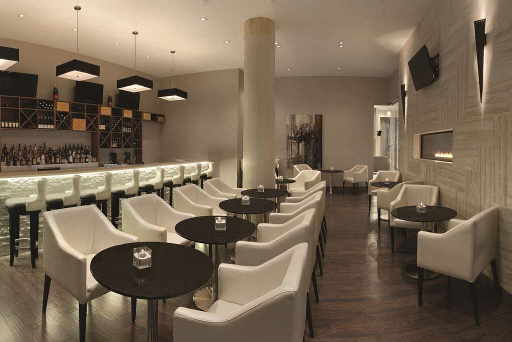 Sfeervolle hotelbar met een lange bar met flessen, ronde tafeltjes met witte leren stoelen en een ingebouwde gashaard tegen een stenen muur