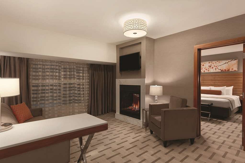 Ruime hotelsuite met een zithoek met fauteuil, een ingebouwde gashaard onder een televisie en een doorkijk naar een aparte slaapkamer met kingsize bed