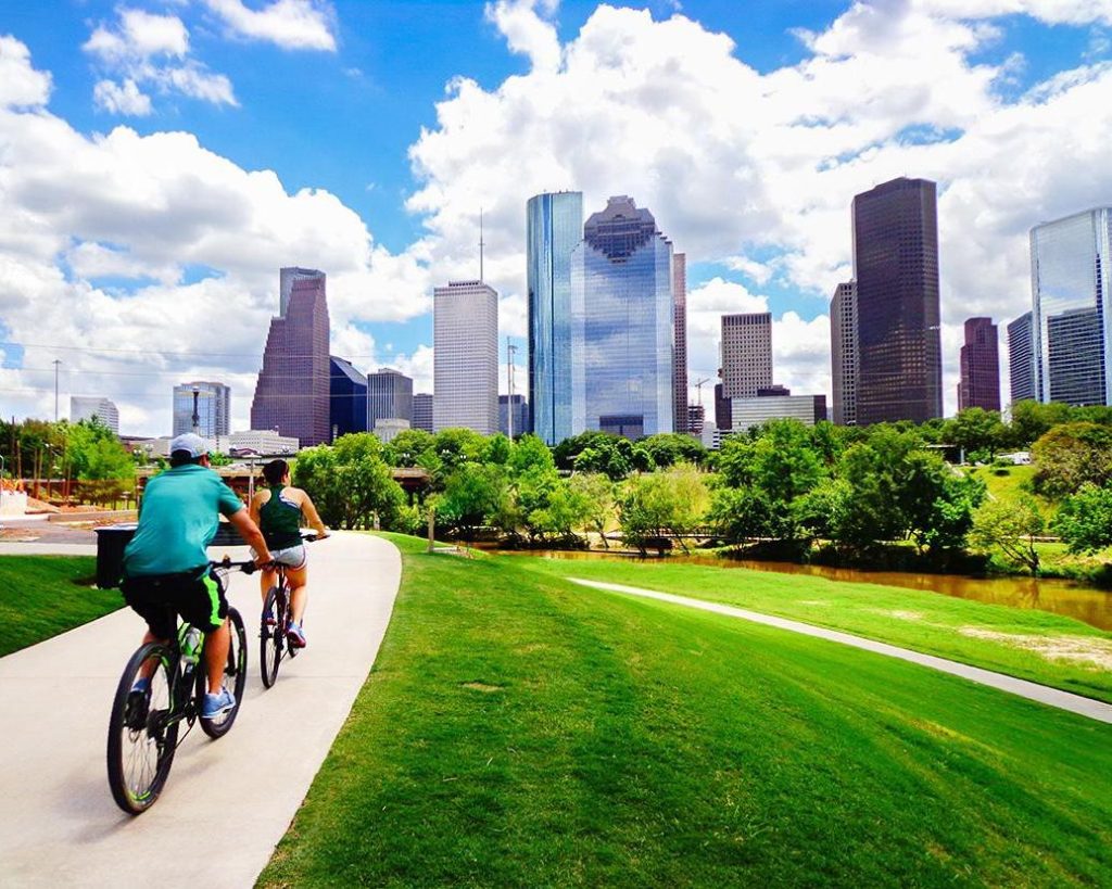 Twee fietsers rijden over een pad door een groen park met de wolkenkrabbers van Houston op de achtergrond onder een blauwe lucht