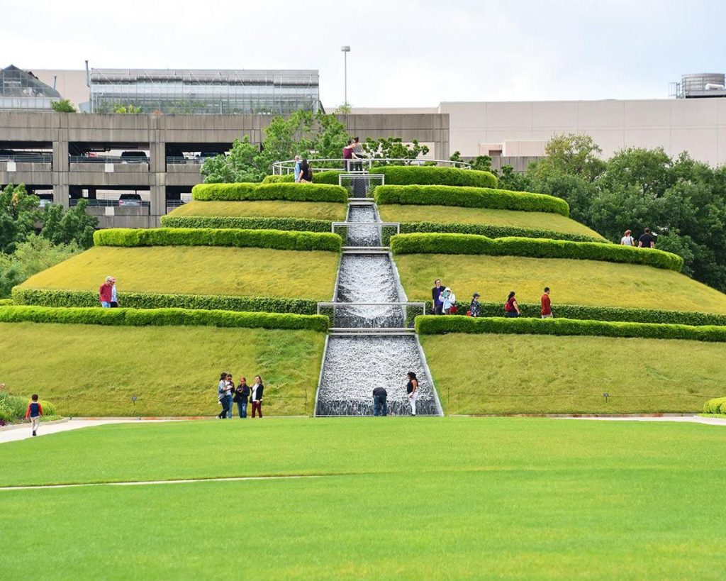 Groene, piramidevormige heuvel met een getrapte waterval waar bezoekers overheen lopen in Discovery Green park in Houston