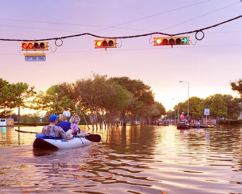 Mensen peddelen in opblaasbootjes over een ondergelopen straat met verkeerslichten die boven het water hangen bij zonsondergang