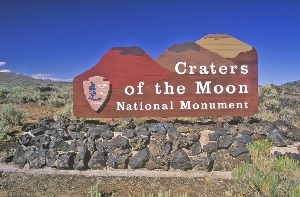 Houten informatiebord van Craters of the Moon National Monument staat op een muurtje van lavastenen onder een strakblauwe hemel