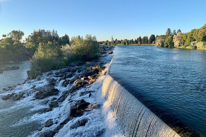 Water stroomt glinsterend over een lange stuwdam in de rivier, met bomen langs de oevers en een gebouw met koepel in de verte