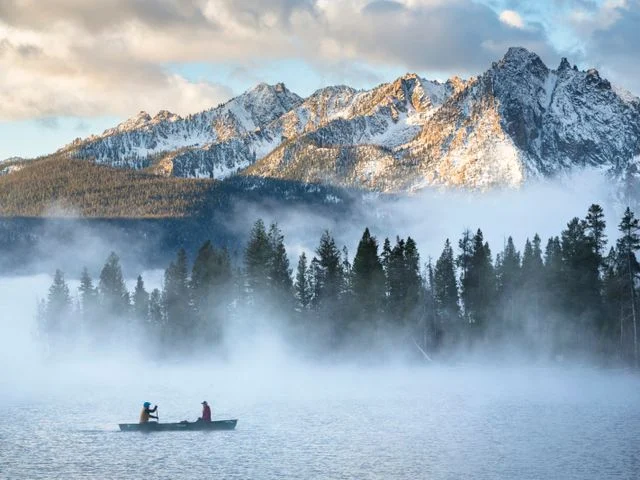 Twee mensen kanoën over een mistig meer met besneeuwde bergpieken en dennenbos op de achtergrond in Idaho