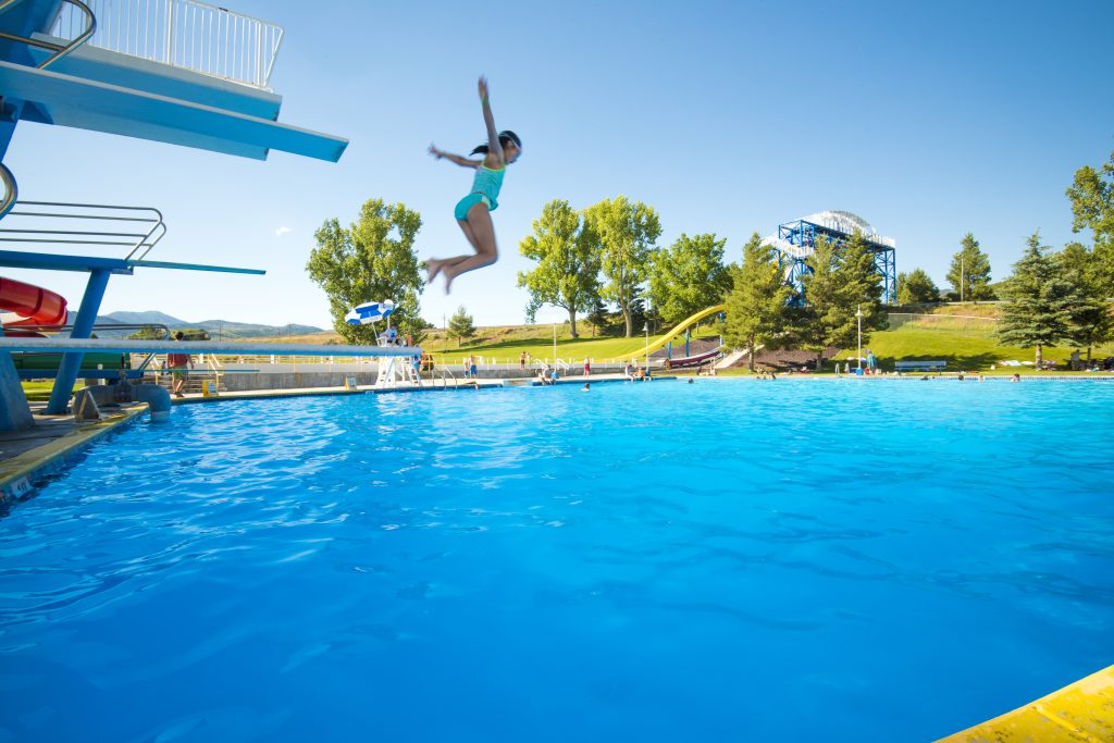 Meisje in turquoise badpak maakt een sprong vanaf een hoge duikplank boven een helderblauw zwembad met glijbanen en heuvels op de achtergrond