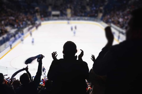 Silhouetten van juichende fans met opgeheven handen en een sjaal op de tribune met de ijsbaan op de achtergrond