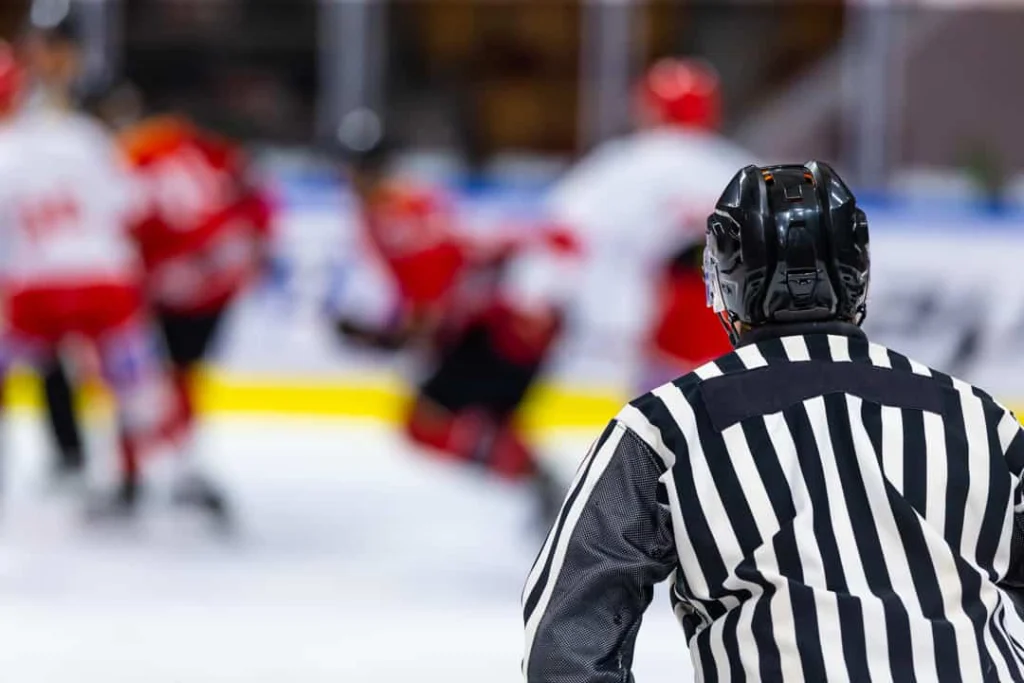 Scheidsrechter in zwart wit gestreept shirt en helm kijkt naar spelers die vervaagd op de achtergrond duelleren