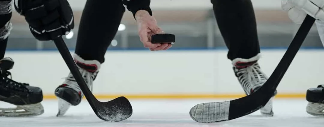 Close-up van twee ijshockeysticks en een zwarte puck tussen de benen en schaatsen van twee spelers op het ijs