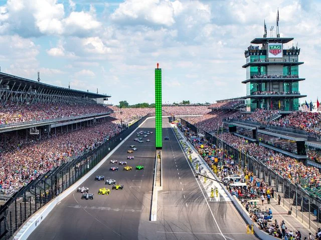 Racewagens rijden in formatie over het rechte stuk van de Indianapolis Motor Speedway tussen volle tribunes met toeschouwers