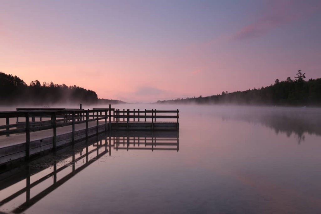 Houten steiger die het spiegelgladde water in loopt, met opstijgende mist en een roze lucht bij zonsopgang