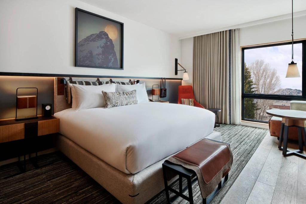 Hotelkamer met een wit bed, een leren poef, een rode fauteuil en een groot raam met uitzicht op bergen en bomen