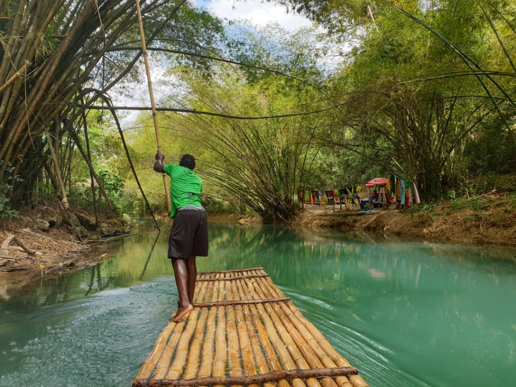 Een man in groen shirt vaart een bamboevlot over groen water met kleurrijke kraampjes aan de oever op de achtergrond in Jamaica
