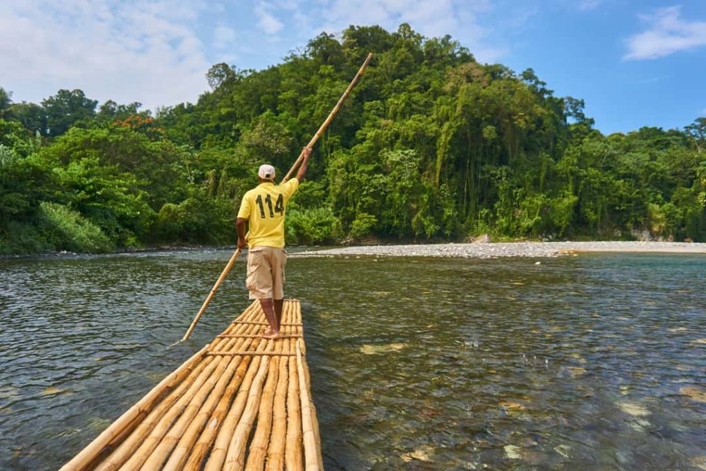 Een man met het nummer 114 op zijn shirt poolt een bamboevlot over ondiep rivierwater langs een jungleoever in Jamaica