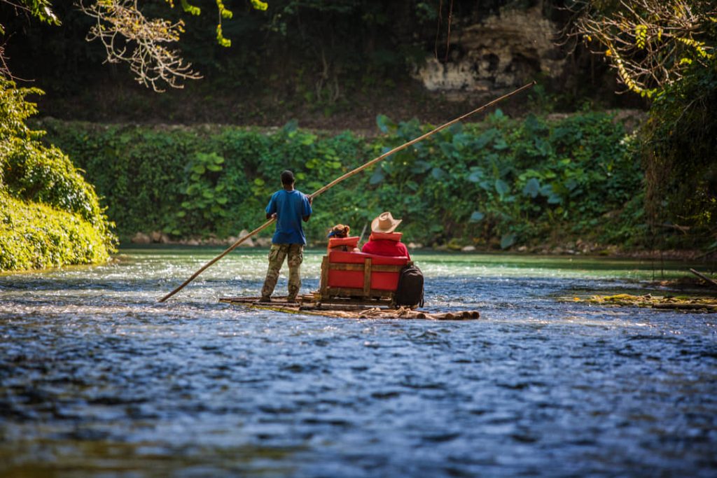 Een gids poolt een bamboevlot met twee passagiers op rode kussens over een ondiepe rivier tussen groene oevers in Jamaica