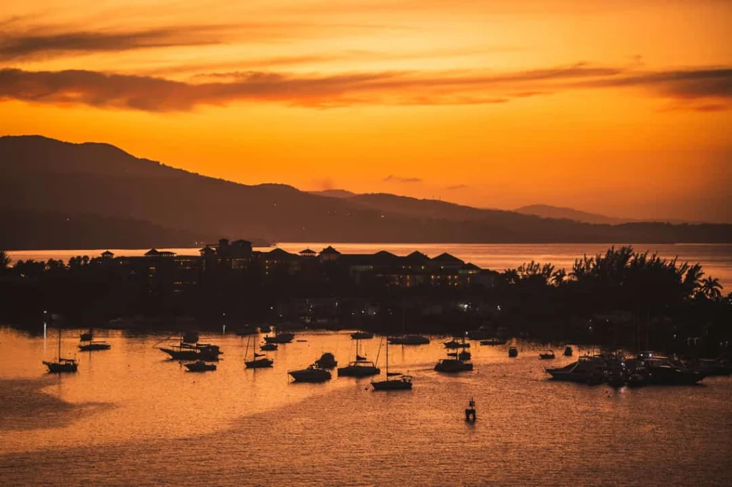 Tientallen bootjes liggen afgemeerd in een baai terwijl de lucht oranje kleurt bij zonsondergang met bergen op de achtergrond