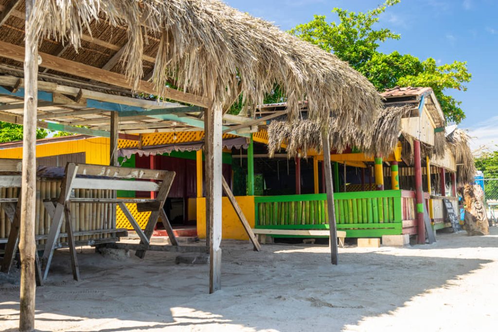 Een kleurrijke strandbar met rieten dak en groen-geel geschilderde houten wanden op een zandstrand in Jamaica