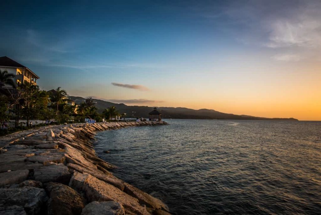 Een stenen kademuur langs de kust van Jamaica met resortgebouwen en palmbomen tijdens een kleurrijke zonsondergang over zee