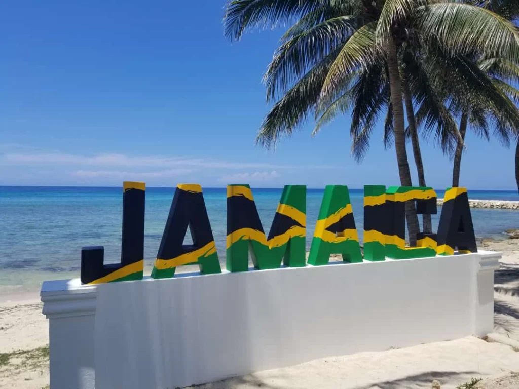 Grote gekleurde letters met de tekst Jamaica op een wit strand met palmbomen en helderblauw water op de achtergrond