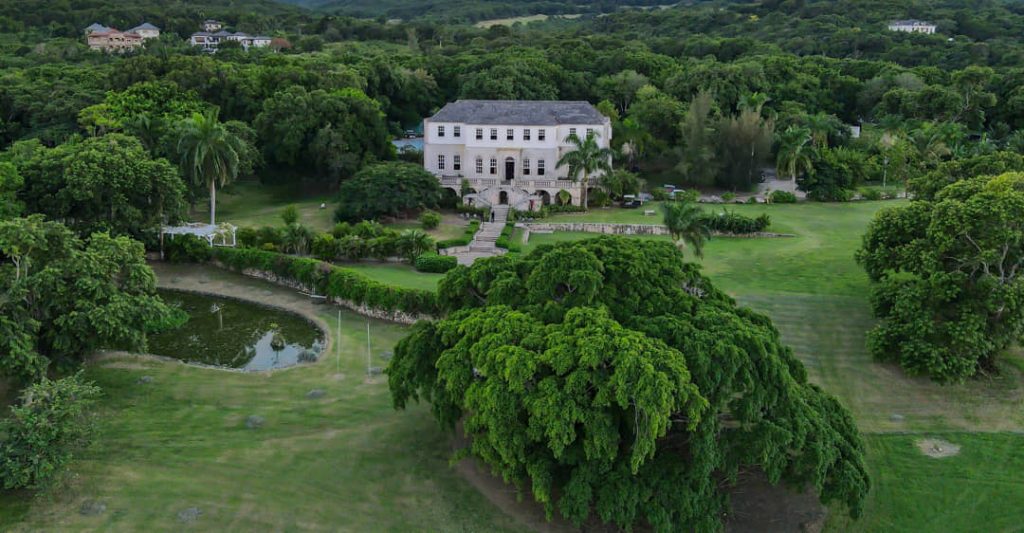 Luchtfoto van een wit koloniaal plantagehuis omringd door bomen, gazon en een ronde vijver in Jamaica
