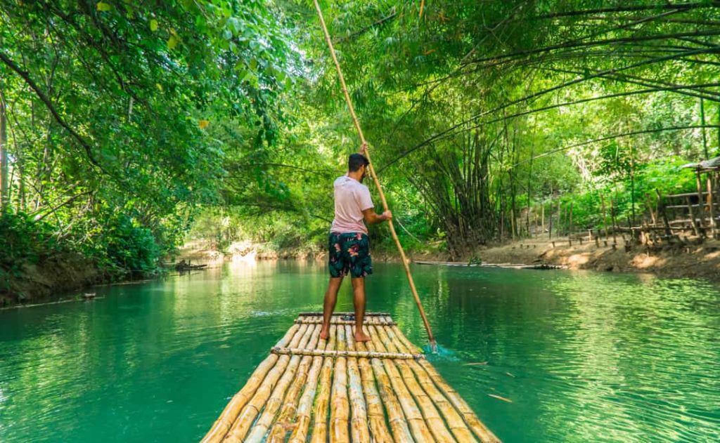 Een man vaart met een lange stok een bamboevlot voort over een groen rivier tussen dichte jungle en bamboe in Jamaica