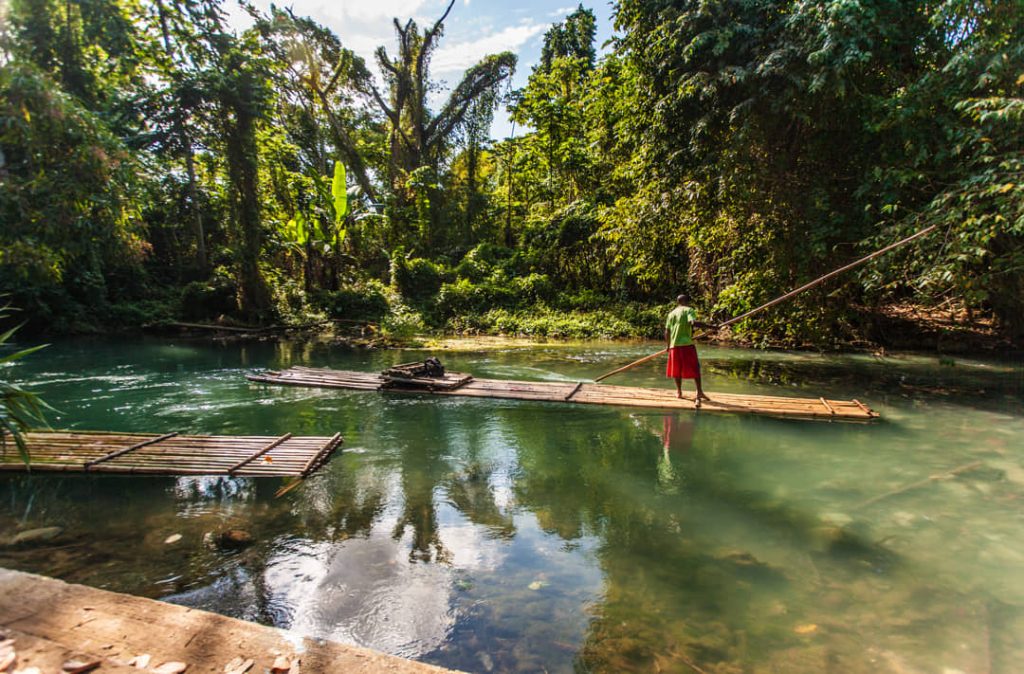 Een man in rode korte broek loopt over een bamboevlot met bagage terwijl hij het met een stok voortduwt over ondiep water in Jamaica