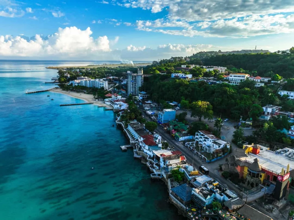 Luchtfoto van een kustplaats met witte gebouwen, een hoge toren en een strand langs helderblauw water in Montego Bay