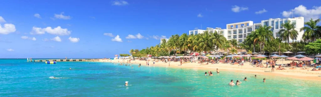 Een druk zandstrand met parasols en zwemmers ligt voor een groot wit resortgebouw omzoomd door palmbomen in Montego Bay