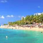 Een druk zandstrand met parasols en zwemmers ligt voor een groot wit resortgebouw omzoomd door palmbomen in Montego Bay