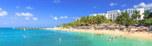 Een druk zandstrand met parasols en zwemmers ligt voor een groot wit resortgebouw omzoomd door palmbomen in Montego Bay