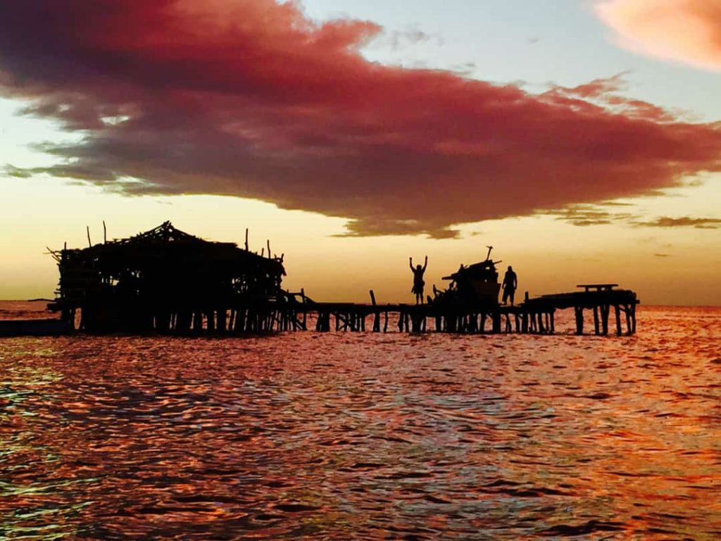 Silhouet van een houten strandbar op palen in zee met mensen erop, tegen een dramatische rode zonsondergang