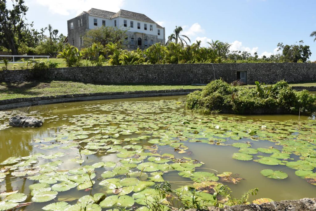 Een wit koloniaal plantagehuis staat op een heuvel achter een vijver vol waterlelies in Jamaica