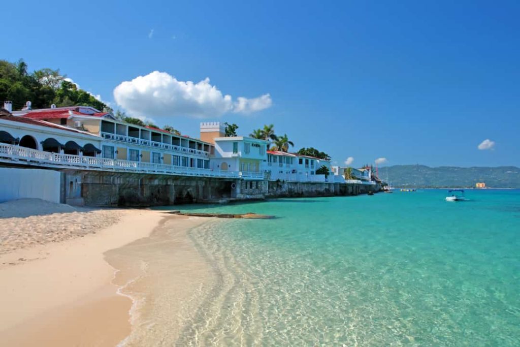 Wit en oranje koloniale hotelgebouwen staan direct aan een smal zandstrand met helder turkoois water in Jamaica
