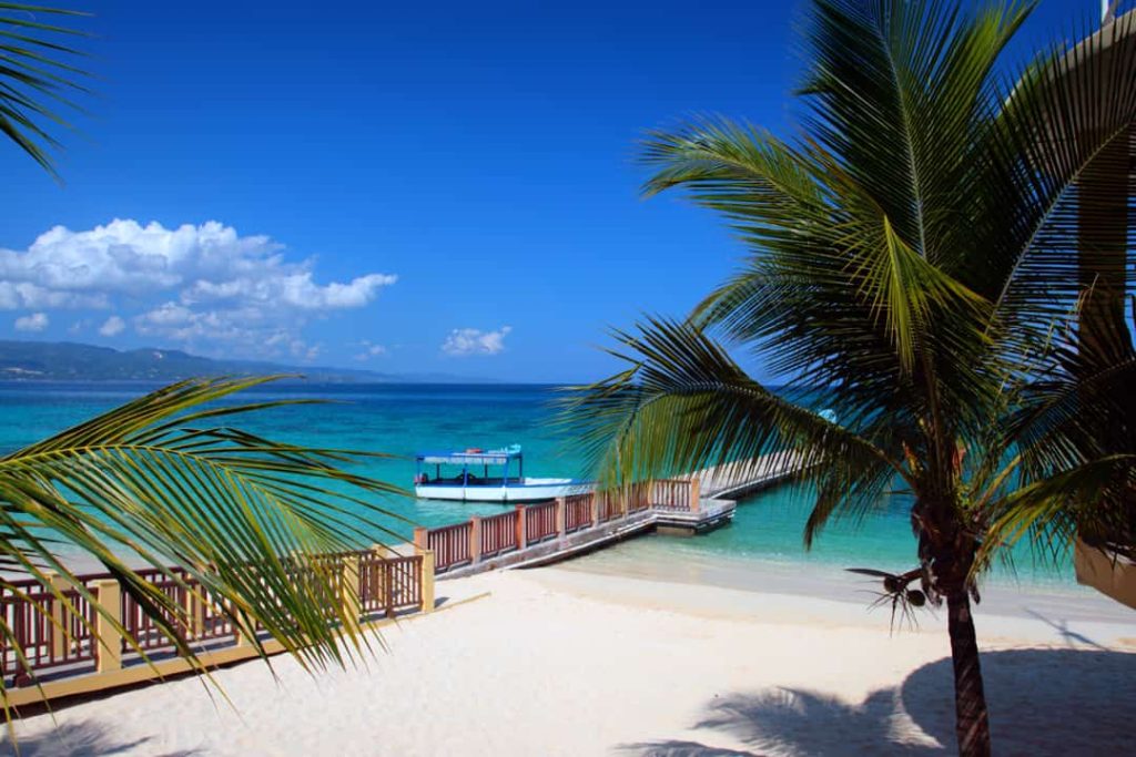 Een houten steiger met een boot ligt aan een wit zandstrand tussen palmbomen onder een blauwe lucht in Jamaica