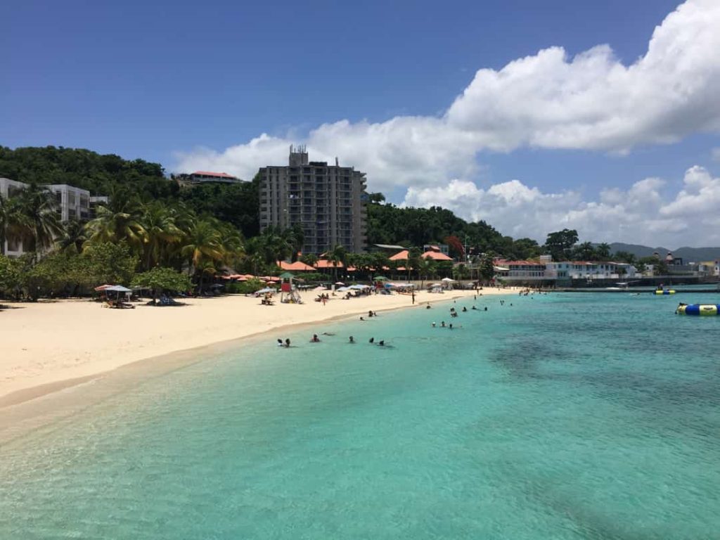 Mensen zwemmen in helder turkoois water voor een wit zandstrand met een hoog hotelgebouw op de achtergrond in Jamaica