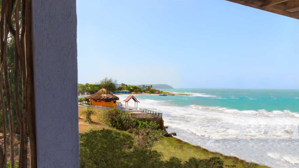 Uitzicht vanaf een overdekt balkon over een tropisch strand met golven, een strohut en een baai op de achtergrond