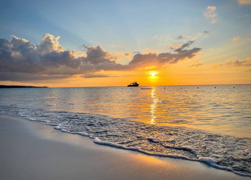 Zachte golven spoelen op het strand terwijl de zon oranje kleuren werpt over zee met een boot op de achtergrond in Jamaica