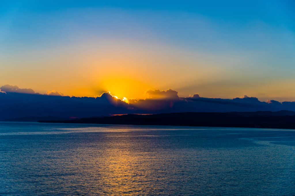 De zon zakt achter donkere heuvels en werpt een goudgele gloed over rustig zeewater in Jamaica