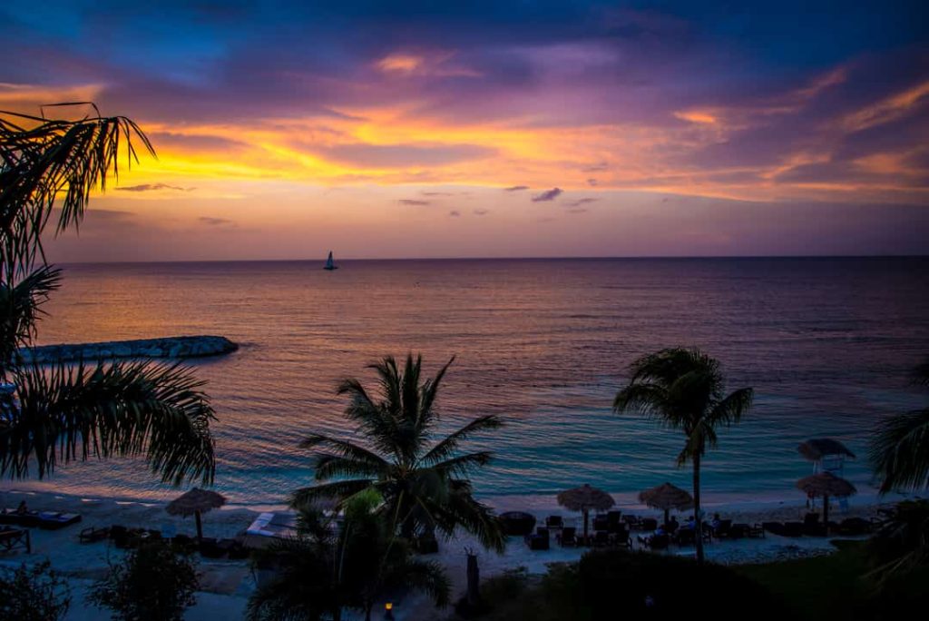 Silhouetten van palmbomen tegen een paarse en oranje avondlucht boven een rustige zee in Jamaica