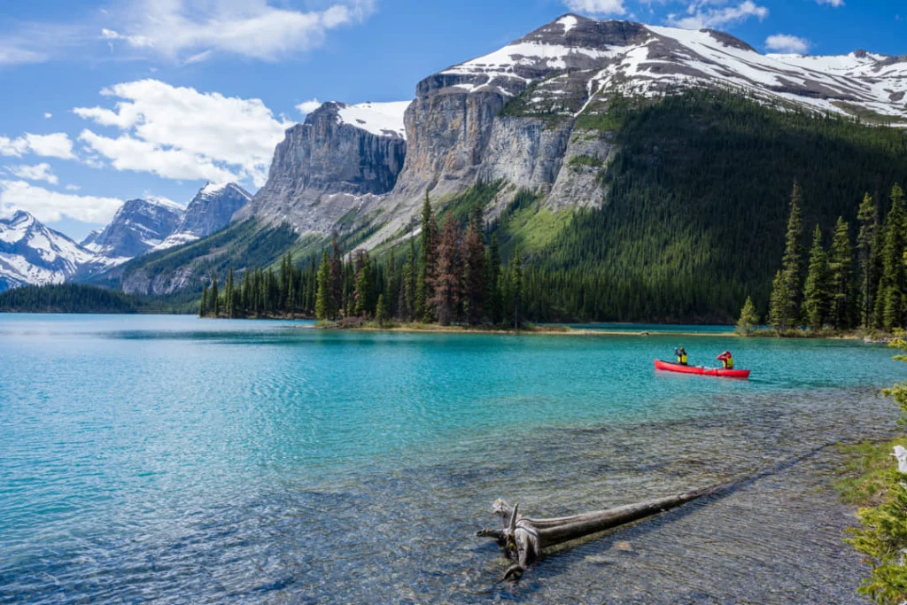 Rode kano op turquoise water van Maligne Lake met een boomstam op de voorgrond en besneeuwde bergen op de achtergrond