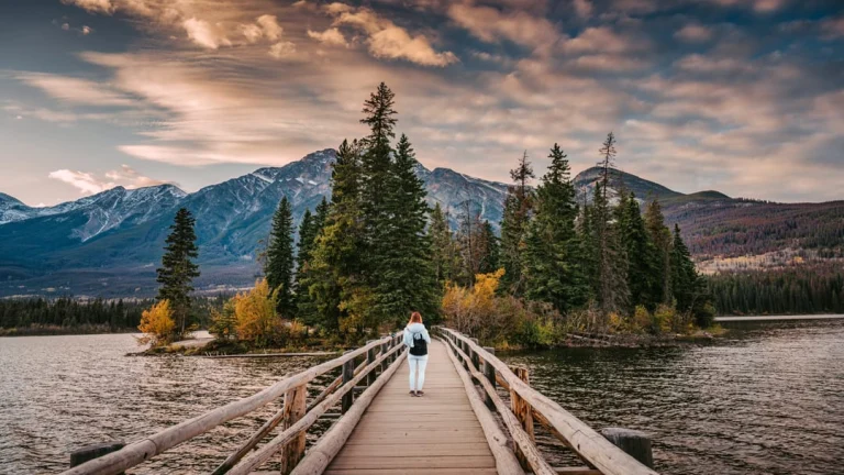 Een vrouw met rugzak loopt over een houten brug naar een klein bosrijk eilandje in Pyramid Lake, met besneeuwde bergen op de achtergrond in Jasper National Park
