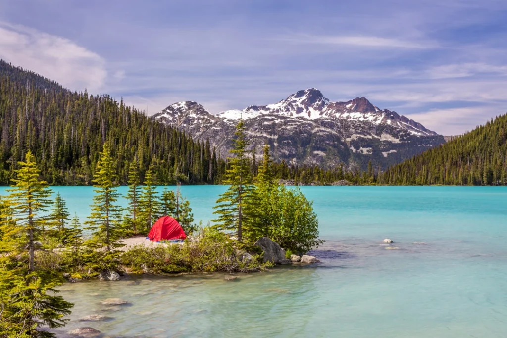 Felle turkooizen kleur van Joffre Lake met een rode kampeertent tussen dennen aan de oever en besneeuwde bergen op de achtergrond in British Columbia