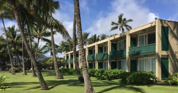 Laag hotelgebouw met groene balkons omringd door palmbomen en een grasveld op Kauai