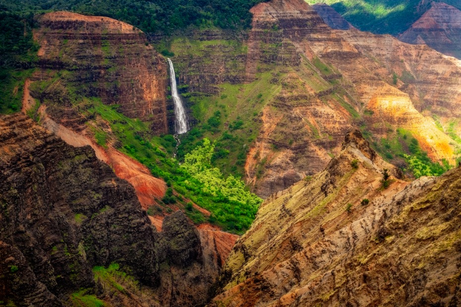 Smalle waterval die tussen roodbruine rotswanden en groene begroeiing naar beneden valt in Waimea Canyon op Kauai