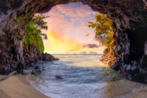 Rotsachtige zeegrot met begroeiing die uitkijkt op een strand en een kleurrijke zonsondergang boven zee op Kauai