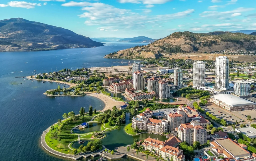 Luchtfoto van het centrum van Kelowna met hoge appartementsgebouwen, een strand en een jachthaven aan het Okanagan-meer, omringd door heuvels