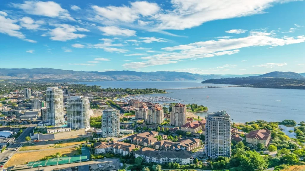 Luchtfoto van Kelowna met hoge appartementengebouwen aan het meer, omringd door bergen onder een blauwe lucht